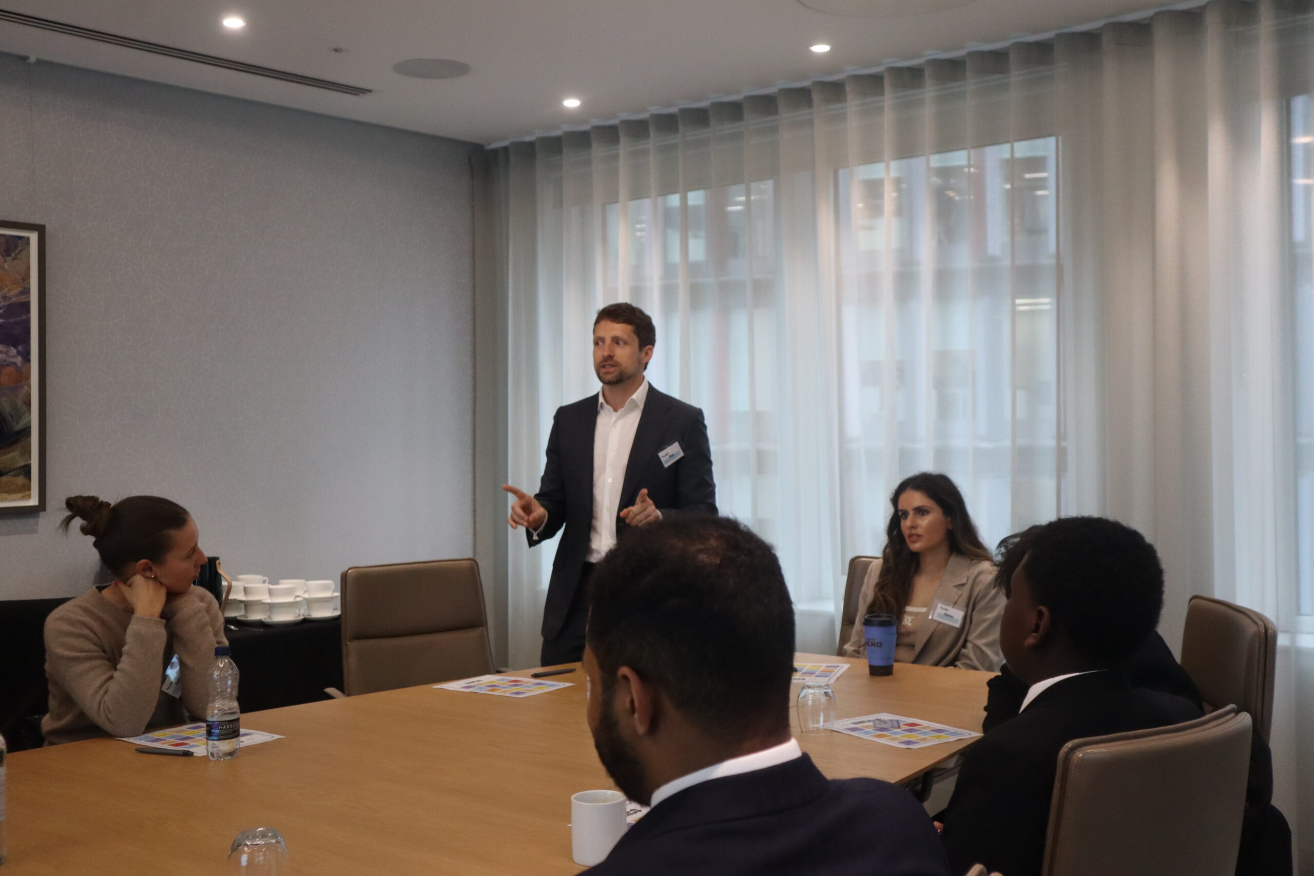 Man stood at next to a table, giving a presentation to a group of students.